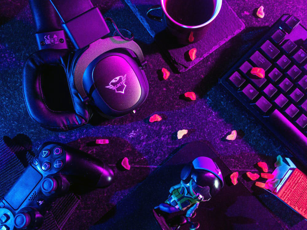 a set of headphones, gaming controllers, a computer keyboard, some dispersed snacks, and a mug of coffee on a desk