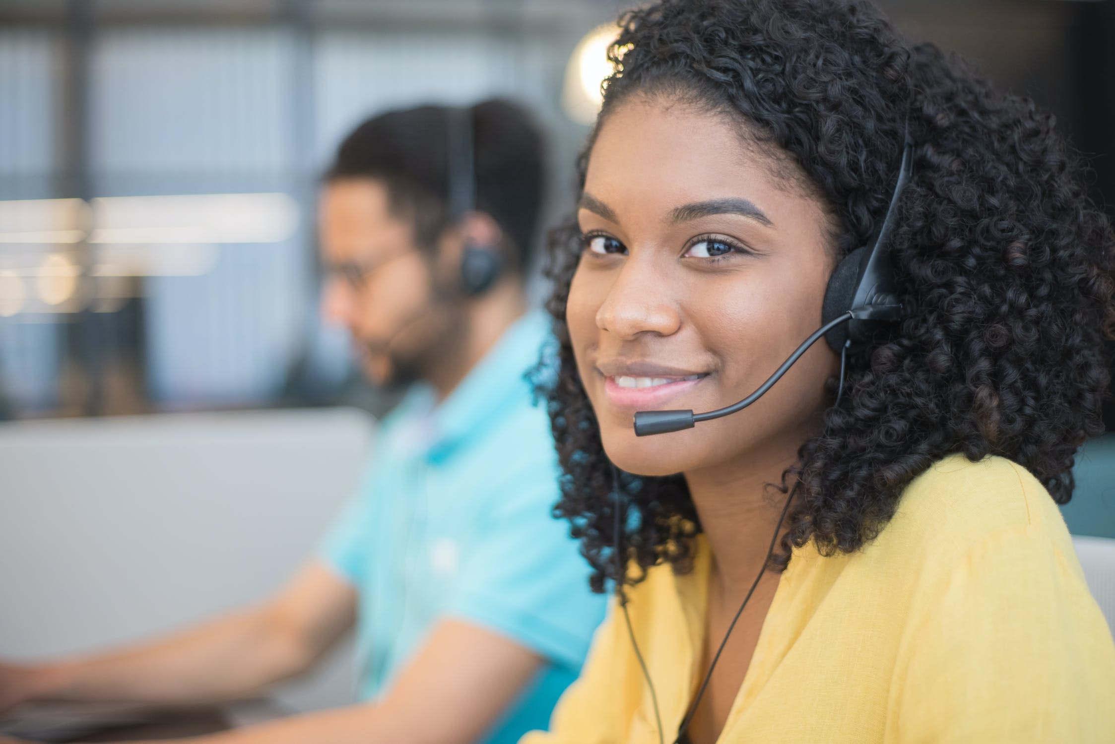 a call center woman with a headset wearing a yellow blouse with another call center employee in the background