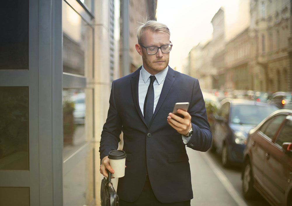 man in a suit looking at his phone in one hand and a to-go coffee cup in his other hand