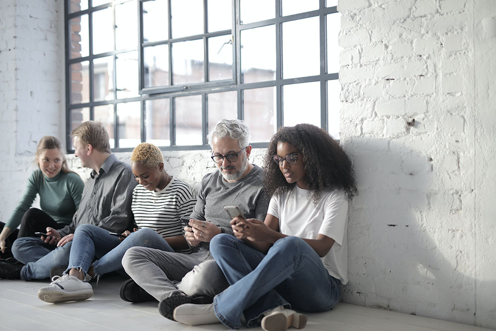 five people of varying ages and races sitting next to each other in front of a window looking at their phones
