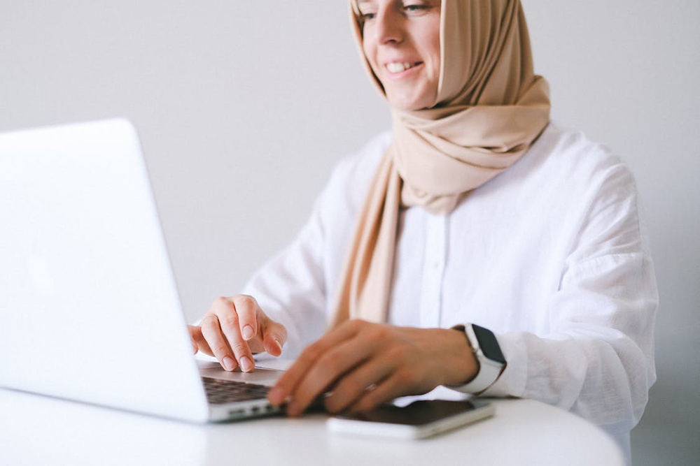 woman wearing a hijab on a laptop with a phone on the desk nxt to her