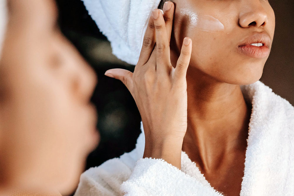 a woman in a bathrobe and a towel in her hair while applying a skin care product on her face while looking in the mirror