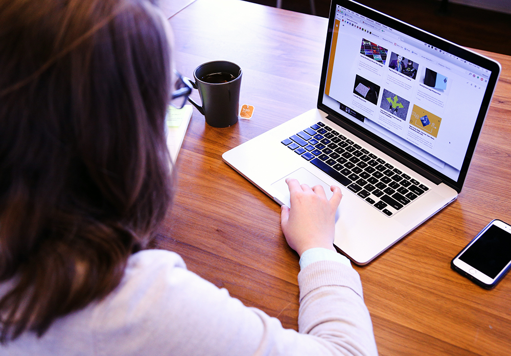 a woman looking at a website on her laptop with a mug of tea on the left and an iphone on the right